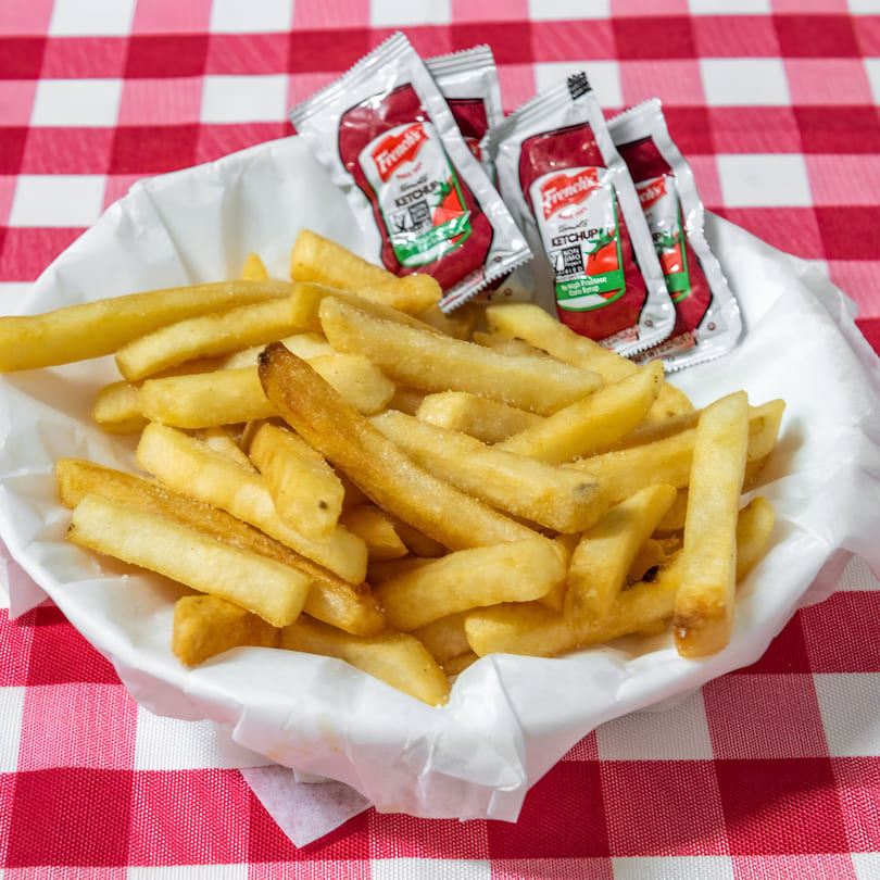 A white paper basket filled with golden french fries and several ketchup packets on a red and white checkered tablecloth.