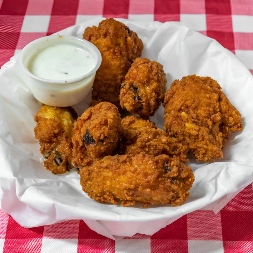 A basket of crispy fried chicken wings with a side of ranch dressing on a red and white checkered tablecloth.