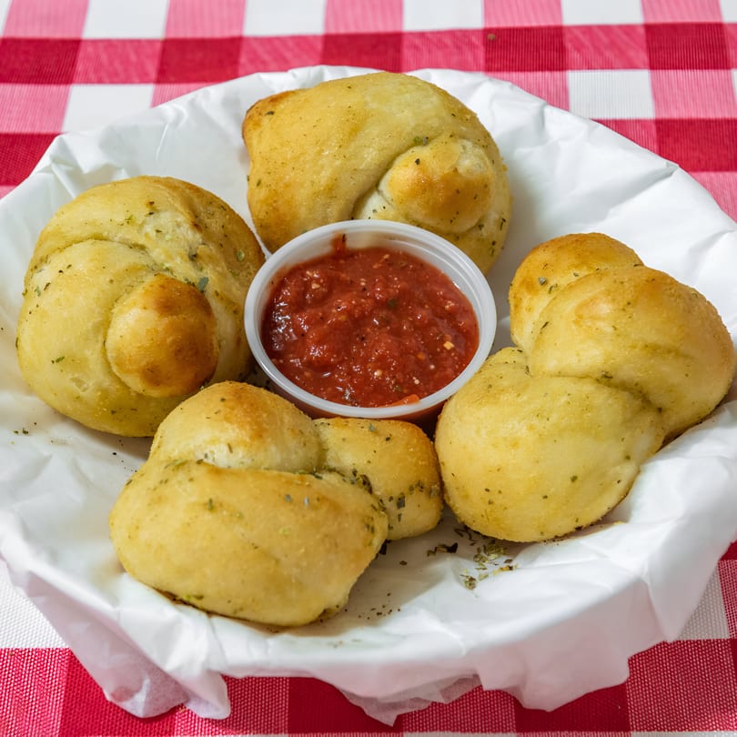 Golden garlic knots with herbs and a side of marinara sauce in a white basket on a red and white checkered tablecloth.