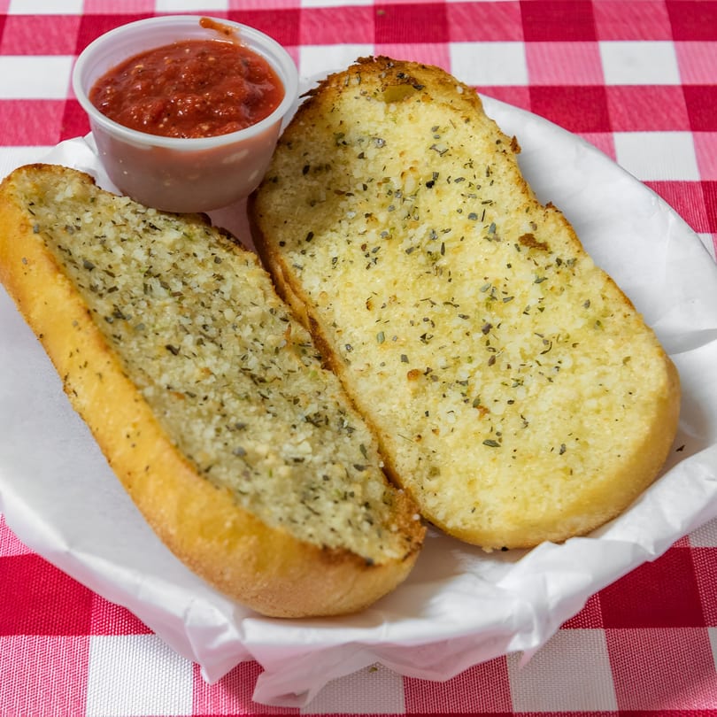 Two slices of garlic bread with herbs and a side of marinara sauce on a red and white checkered tablecloth.