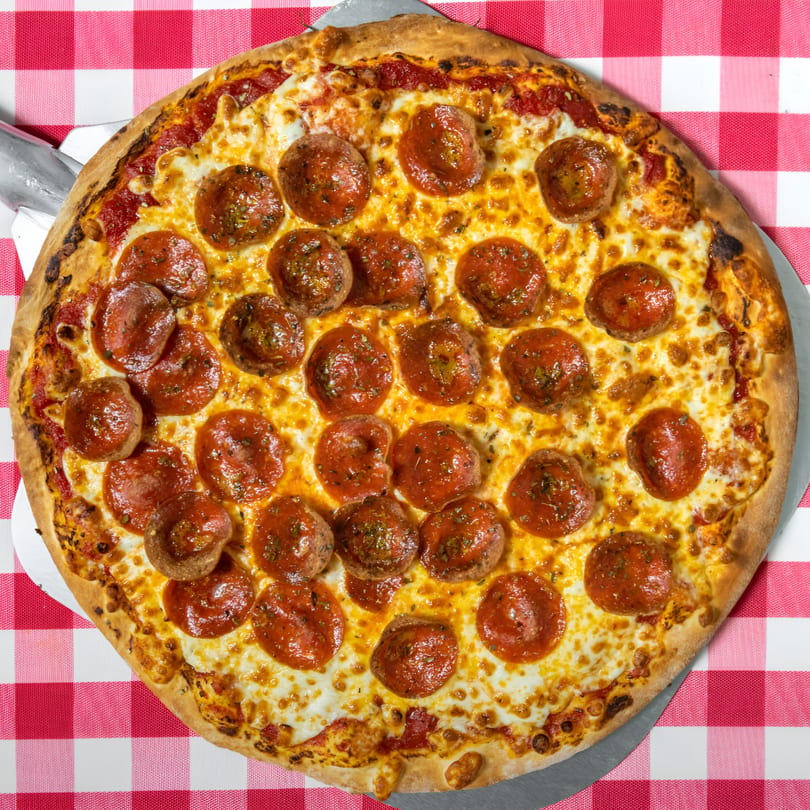 A whole pepperoni pizza with extra cheese and meatballs on a red and white checkered tablecloth.